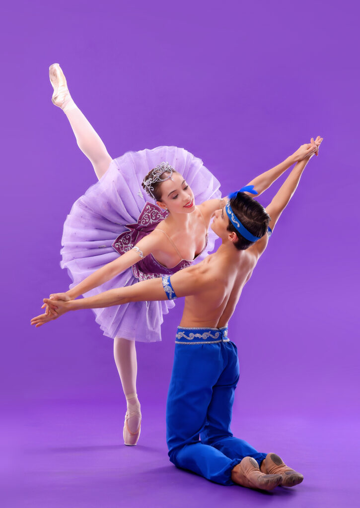 Against a bright purple backdrop, Margarota Fernandes and Antonio Casalinho do a partnered pose. Casalinho, his bac to the camera, kneels on the ground on both knees and spreads his arms wide to match Fernandes as they join hands. She leans towards him in an arabesque penché, looking into his eyes. Her costuem is a purple pancake tutu and tiara, with pink tights and pointe shoes, while he wears bright blue harem pants, blue arm bands and a feathered headband.