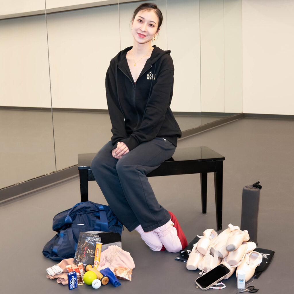 Ye Feifei sits on a piano bench with her dance-bag contents arranged on the floor on either side of her. She is wearing a black hoodie, black exercise pants, and pink and red warm-up boots. Her hands are in her lap, and she is smiling at the camera.
