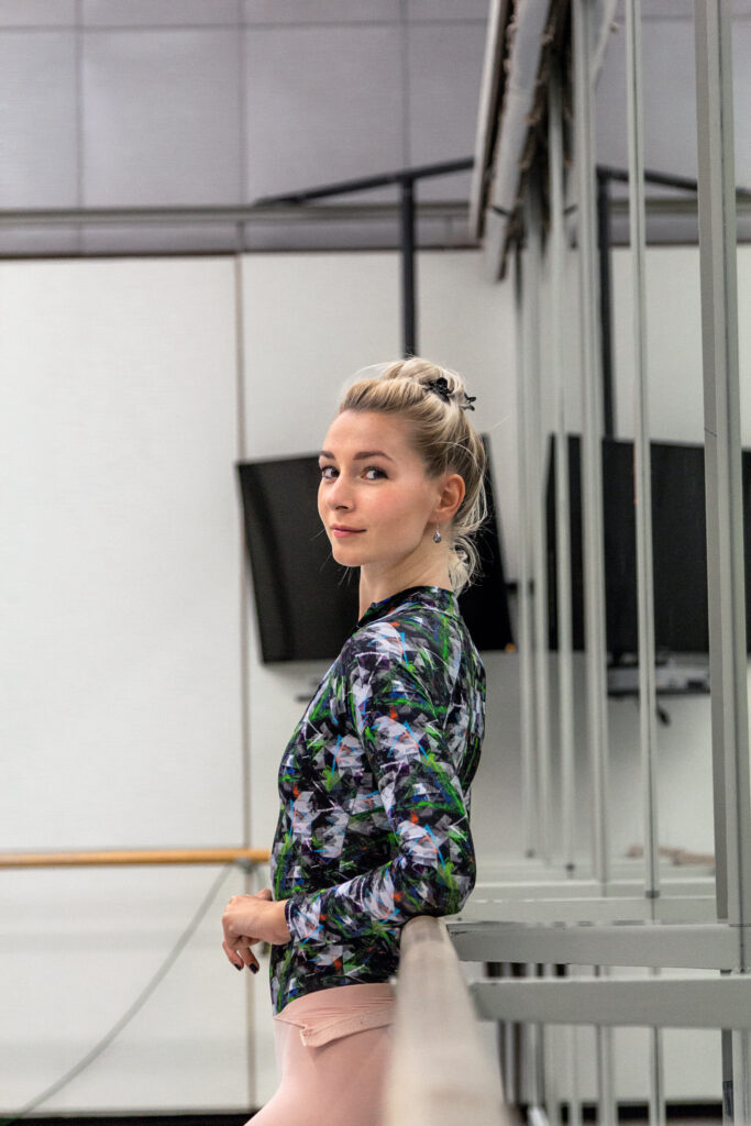 Rebecca Haw, a ballet dancer, is shown in profile leaning against a barre in a large dance studio. She turns her head towards the camera and offers a small, closed mouth smile. She wears a dark, multicolored long-sleeved leotard with pink tights over it. Her blonde hair is in a bun.