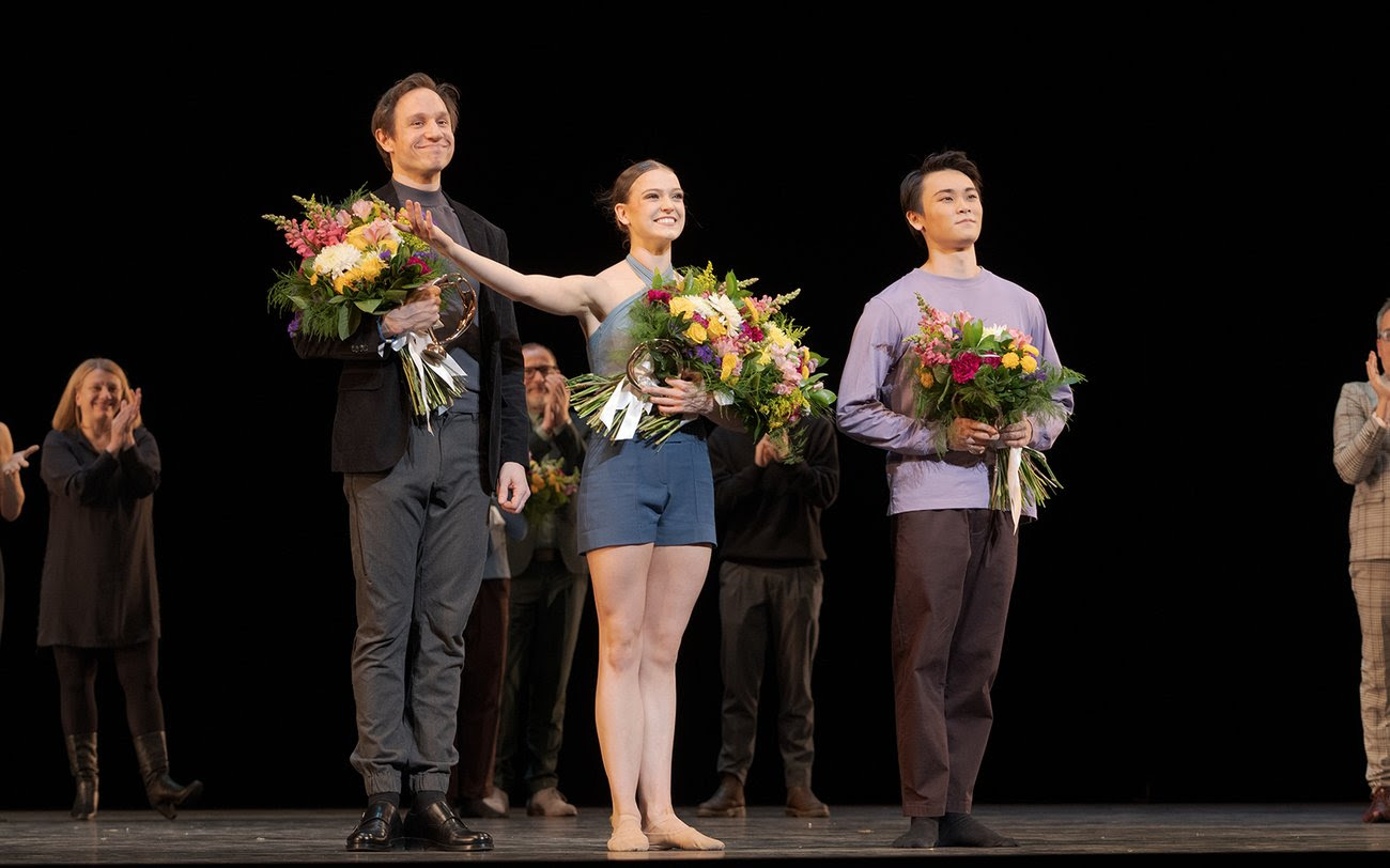 Onstage in front of a black backdrop, Roman Novitzky, Mackenzie Brown and Daichi Ikarashi stand together as prizewinners, each holding a large bouquet of flowers. They stand and smile toward the audience, Brown's right hand extended in thanks. Behind them, artistic directors and other competitors watch and applaud.
