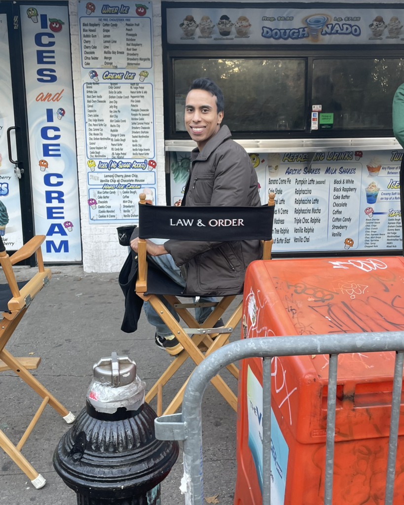Francis Lawrence is photographed from the back sitting in a director's chair with the words Law & Order printed on the back. He turns in the chair to face the camera with a big smile, and wears a brown jacket, jeans and black sneakers. The chair is on a New York City street facing a ice cream shop, with a fire hydrant, metal gate and orange newspaper stand in the foreground.