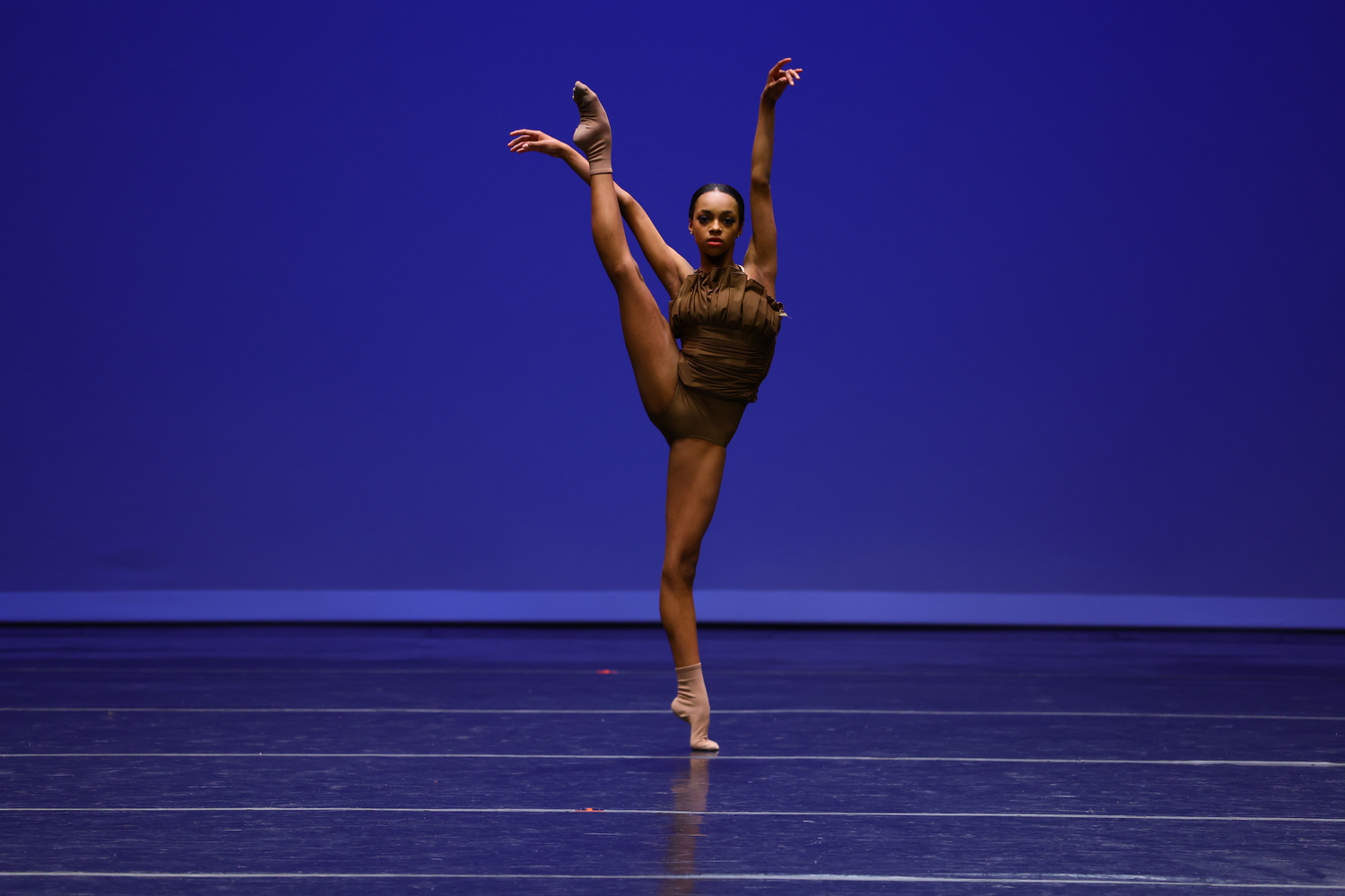 A young ballet student performs a contemporary solo onstage in front of a blue backdrop at YAGP. She wears a brown embellished leotard and tan socks, and does a développé à la seconde with her right leg lifted high. She raises her arms up above her head and looks straight ahead at the audnience.
