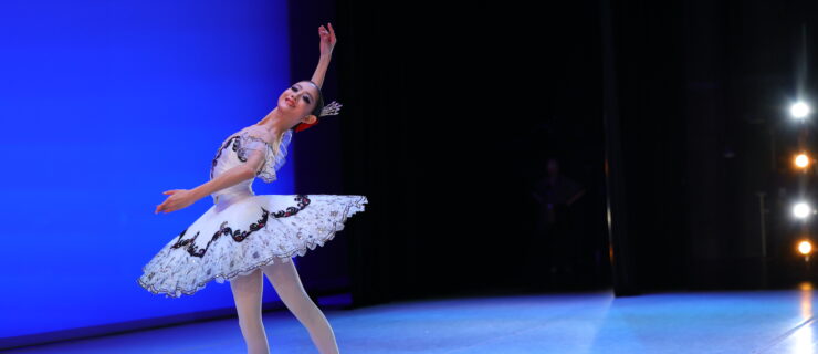 Chae Eun Lee wears a white pancake tutu with black lace trim and poses regally in a tendu derriere facing the upstage right corner onstage during a performance. She holds her arms in fourth position with her right arm raised and leans her upper body back, smiling proudly.