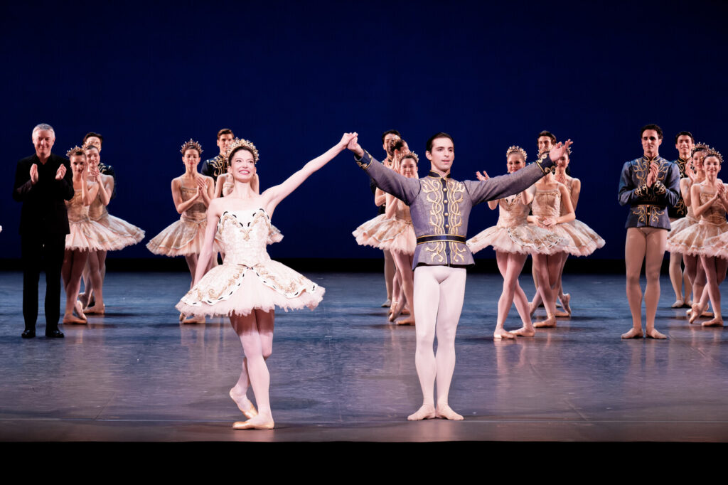 Onstage, a female and a male dancer hold hands and raise their arms in joy as they smile toward the audience, wearing glittering gold costumes. Corps de ballet members smile and clap behind them.