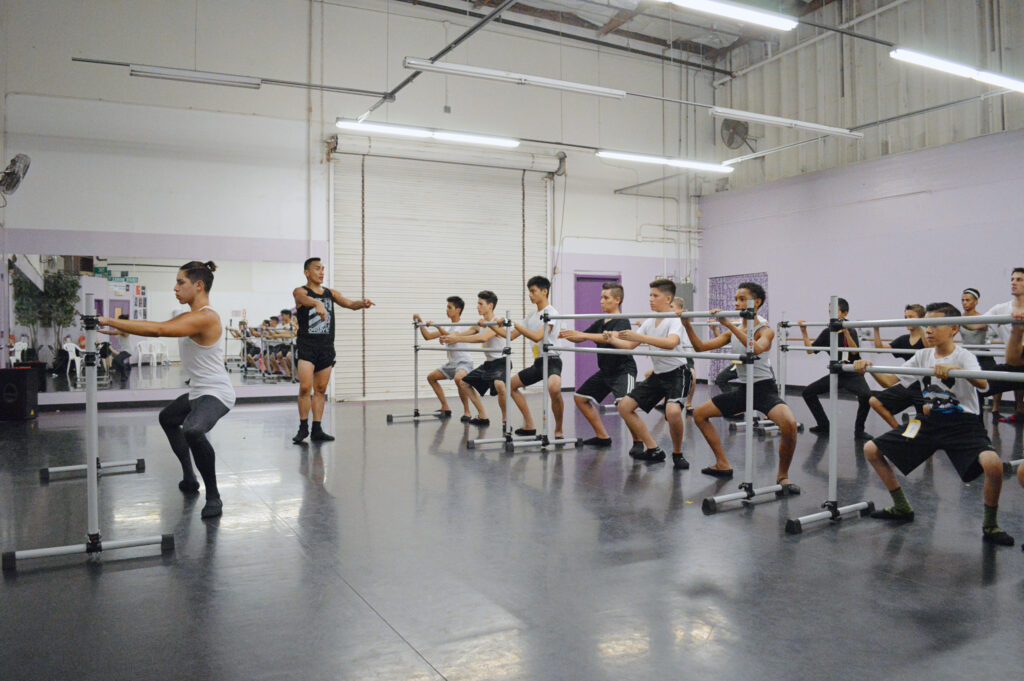 In a large, windowless dance studio, Francisco Gella stands towards teh right side of the room and watches a large class of teenage male students as they do grand plié in second position. Gella—wearing a black sleeveless T-shirt and black shorts, socks and ballet slippers—makes a motion with his hands and corrects the dancers, who wear white or black T-shirts, shorts and ballet slippers.