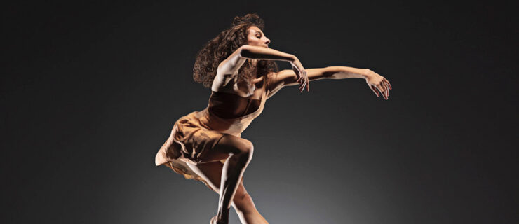 Ilaria Guerra, barefoot and wearing a short brown dance dress and her hair long, jumps up and tucks her right leg slightly underneath her. She bends her upper body forward slightly and extends her curved arms out in front of her, partially covering her face.