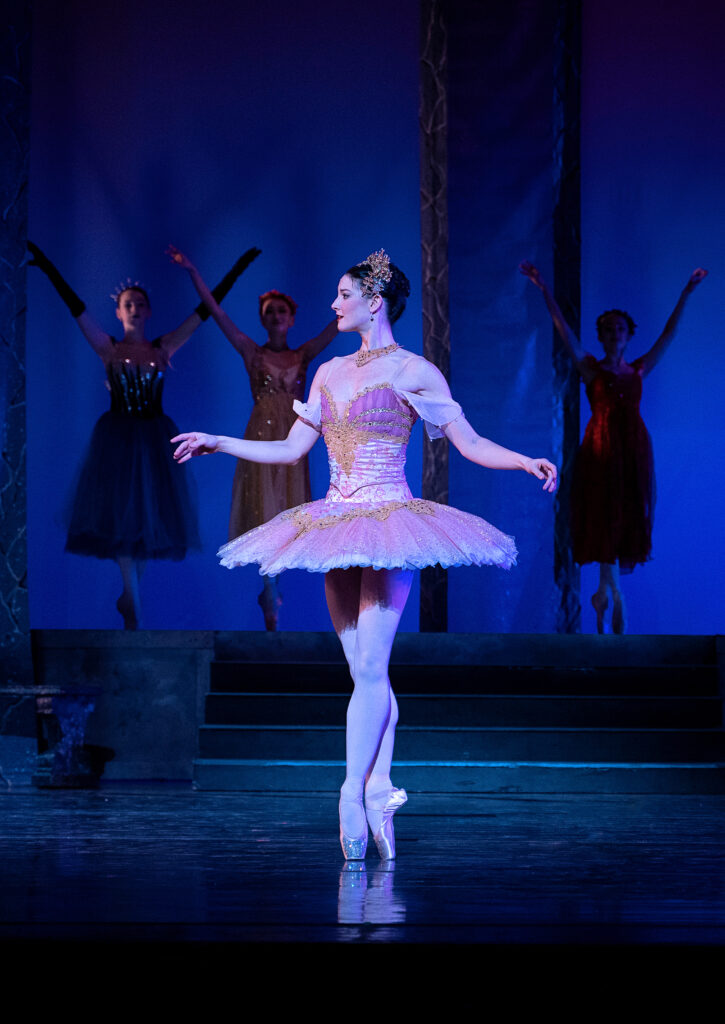 Claire Solis, wearnig a regal pink tutu with gold trim and a gold tiara, stands in profile in sus-sous on pointe, facing stage right. Her arms are slightly relaxed and bent at the elbows, and she is spotlit on a darkened stage. Three dancers in long tutus pose in sus-sous on a short staircase upstage, which is dark, facing the audience.