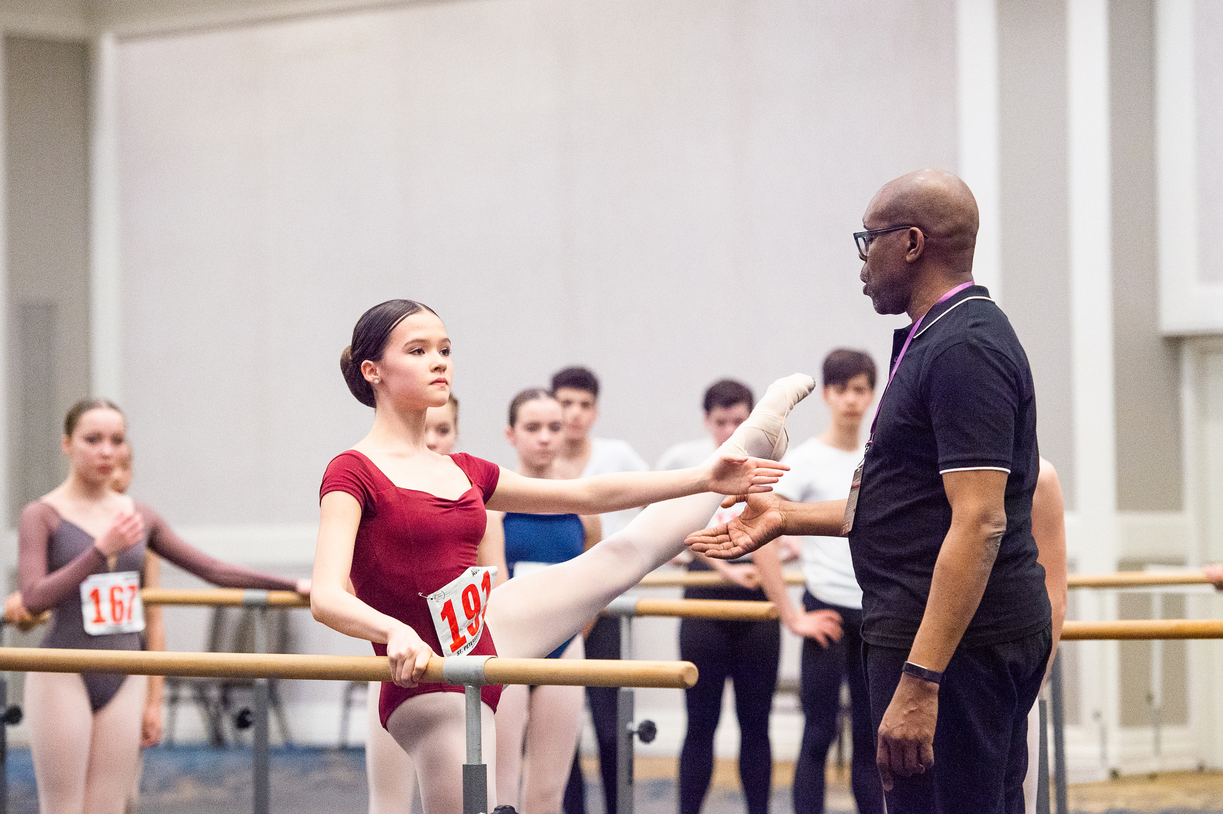 A teenage dancer in a red leotard and wearing the number 191 pinned to her torso does a développé à la seconde at the barrer with her arm in second position. Maurice Brandond Curry, wearing a black polo shirt and black pants, stands in front of her and points to her calf as he gives a acorrection. Other male and females dancers stand around them at the barres and watch.