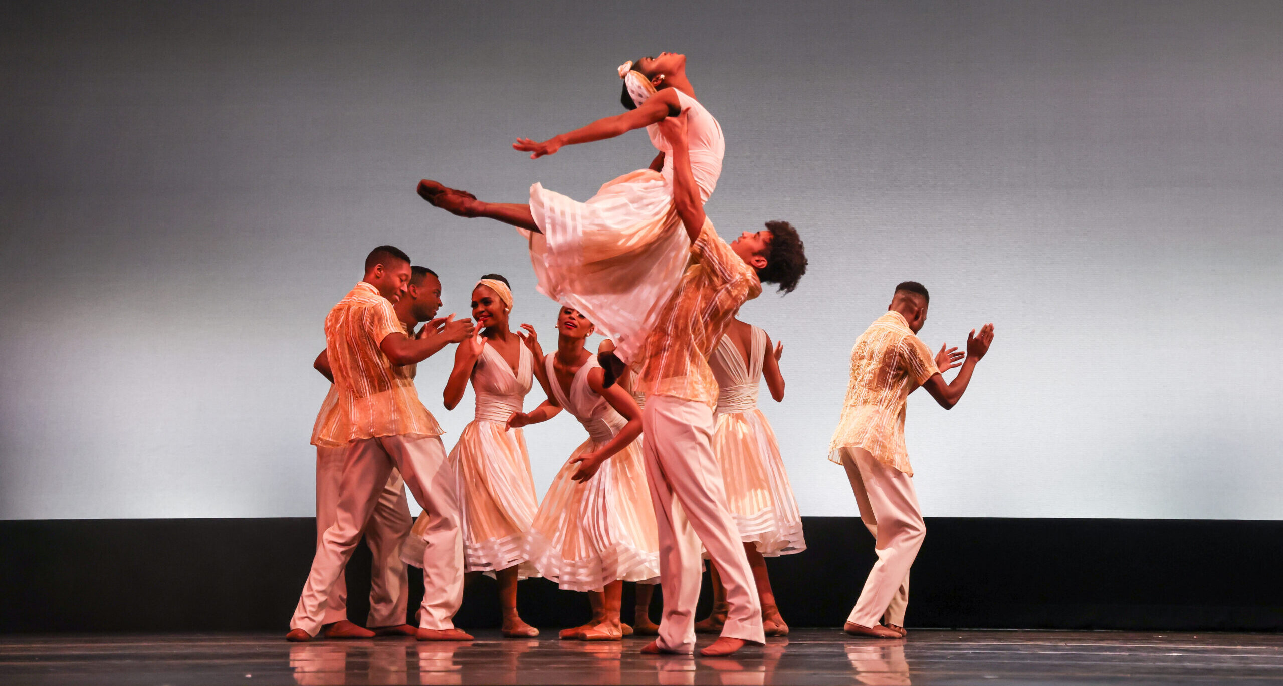 An ensemble of dancers from Dance Theatre of Harlem perform onstage in front of an ombré gray backdrop. In the middle, a female dancer performing as Hazel Scott is lifted and arches back, her head lifted to the sky. The dancers all wear white period-specific dresses and suits.