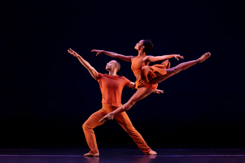 Onstage, a male and female duet dance together in bright orange costumes. In front, the woman flies in a saut de chat, her head lifted above her front arm triumphantly. Behind her, the man lunges forward on his right leg, his arms in a wide fourth position.
