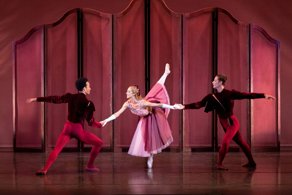 Danielle Brown, costumed in a long pink dress and long white gloves, does a penché in fourth arabesque croise on pointe, hold the hands of two men on either side of her. She faces Ricardo Rhodes, who faces her in a fourth position lunge and holds her right hand, while Daniel Pratt stands to her left, holding her left hand in the same pose but facing the audience. Both men wear wine-colored velvet tunics, red tights and red ballet slippers, and they all dance in front of a dark pink set.