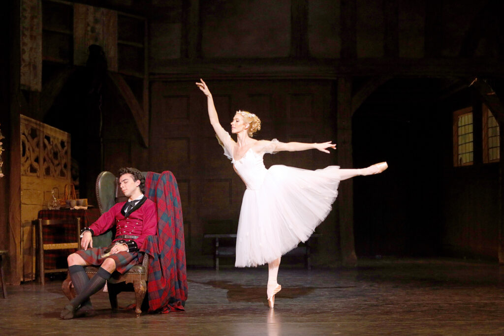 Danielle Brown does a first arabesque on pointe on her right leg towards Maximiliano Iglesias, who sleeps in a large green armchair. She wears a long, white Romantic tutu and wreath of white flowers on her head. Iglesias wears a red jacket, red and green kilt, black knee socks and black ballet slippers. A large red and green tartan hangs over the back of the chair.