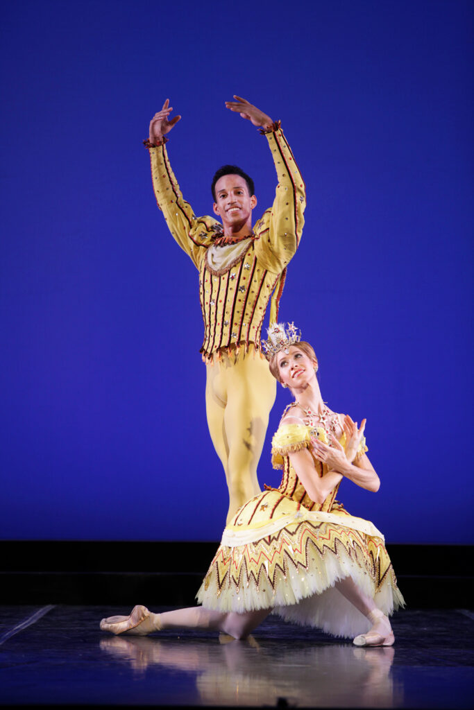 Danielle Brown kneels on her left knee and crosses her wrists in front of her chest, and looks over her right shoulder, smiling broadly. She wears an ornate knee-length yellow tutu and large crown, pink tights and pointe shoes. Behind her, Ricardo Rhodes poses in sus-sous with his left leg in front and his arms in fifth position. He wears a costume of yellow tights and a yellow tunic with puffed long sleeves.
