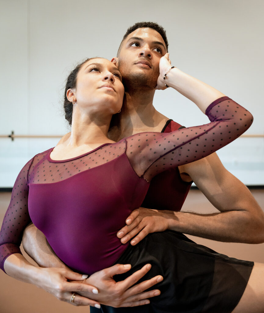A close-up photo shows a woman and a man rehearsing a pas de deux together in a dance studio. The woman, her left leg lifted, leans back against her partner's chest and holds his arm against her stomach, her other arm reaching up to caress his cheek. He steadies her with his other hand on her hip, and they both look into the distance longingly.