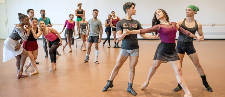 In a large dance studio, Collage Dance Company artists rehearse "Their Eyes Were Watching God." A trio in the front—two men and a woman—perform a partnered traveling step as the corps behind them pose in various poses of gossiping, outrage, and curiosity.