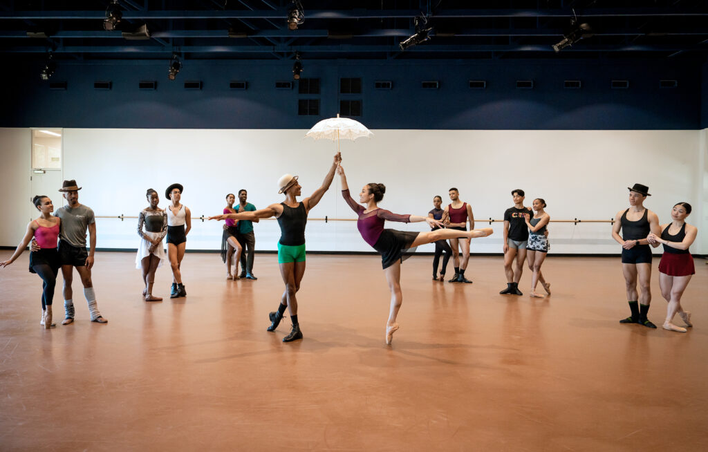 In a large dance studio, Collage Dance Company artists rehearse "Their Eyes Were Watching God." Front and center, a man and a woman dance together, both holding a delicate white parasol. The woman balances on pointe in arabesque and the man stands in B plus as they each hold on to the parasol's handle, which they hold above their heads as they smile at each other. Around them in a semi-circle, six partners of men and women stand in various poses, linking arms or touching to indicate partnership. They watch the center couple. Four of the men, including the one in the middle, wear bowler hats.