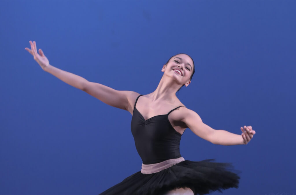 Genesis Robinson, shown from the thighs up, performs onstage in front of a blue backdrop. She wears a black leotard, black practice tutu and pink tights and is shown opening her arms out to the side with a large smile on her face.