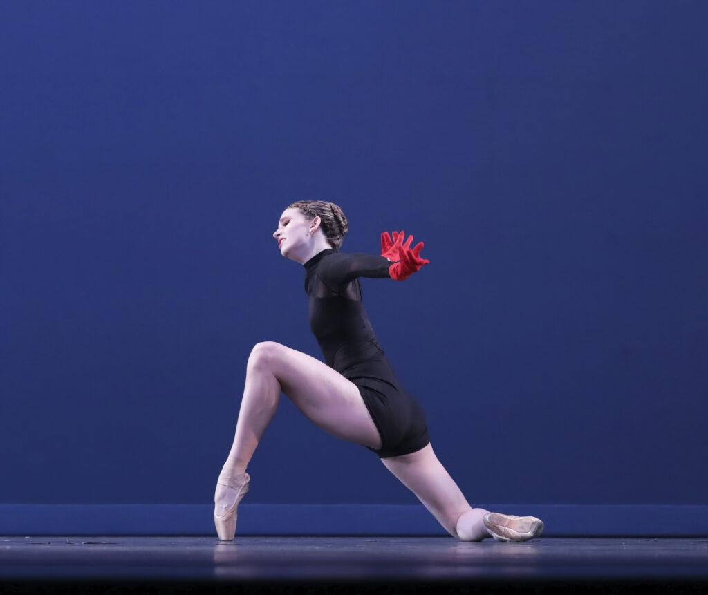 Larenne Taylor performs onstage in front of a blue backdrop. She wears a black biketard, pointe shoes and red gloves. She kneels on her right knee, in profile facing stage right, and props her left foot up onto pointe as she stretches her arms out to the side and slightly behind her. She cocks her head slightly toward her left leg and wears a dramatic expression.