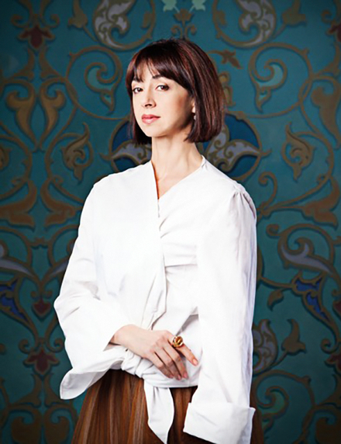 In front of ornate teal and gold arabesque-patterned wallpaper, Nina Ananiashvili, shown hips-up, poses for a portrait in a white cotton blouse and rust-colored trousers. She faces slightly away from the camera and looks into the lens smiling slightly, her right arm folded across her hip.