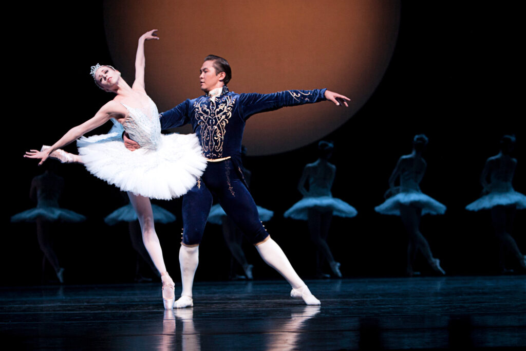 Lit by a spotlight on a dark stage lined by a corps de ballet of swans, a ballerina performing the role of Odette in "Swan Lake" leans back in a supported arabesque, held gently by her partner, who lunges toward her in a tendu a la seconde. He wears a dark blue velvet tunic and tights.