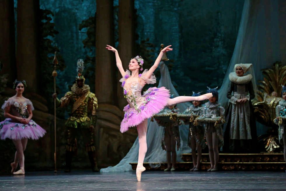 A ballerina in a lilac-colored pancake tutu smiles radiantly as she poses in arabesque onstage, her arms lifted gently in a "V" shape. Behind her, the scene is set for the Prologue of "The Sleeping Beauty."