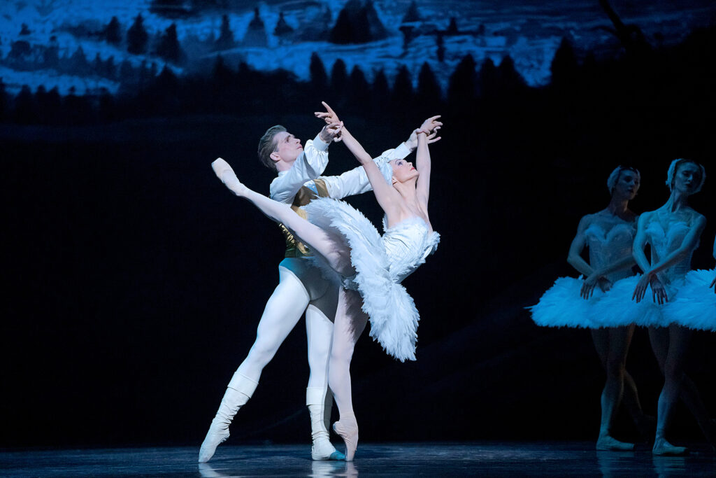 Jurgita Dronina, as Odette in a white feathered tutu, balances on pointe in a high arabesque as she arches backwards. Harrison James, as Siegfried, holds her wrists. Her arms are in a V overhead.