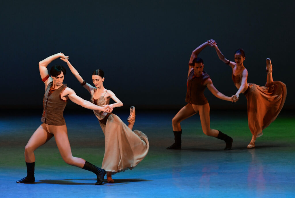 Onstage and lit in dark blue, two pairs of male and female dancers perform a mirrored pas de deux in neutral-colored costumes.