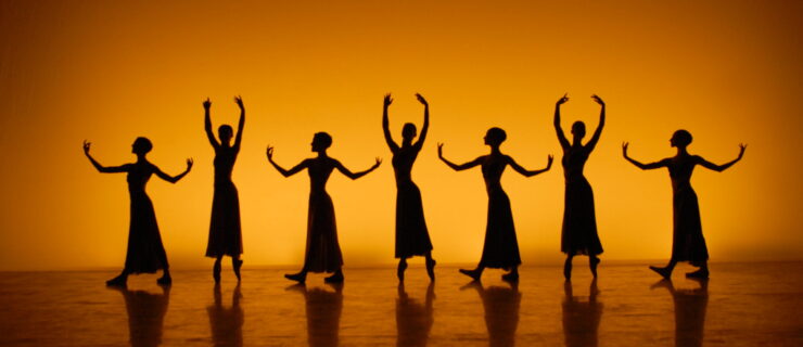 A tableau of female dancer silhouettes line the back of a stage lit with orange in a sunset effect on the backdrop. The women wear skirts and alternate poses every other dancer.
