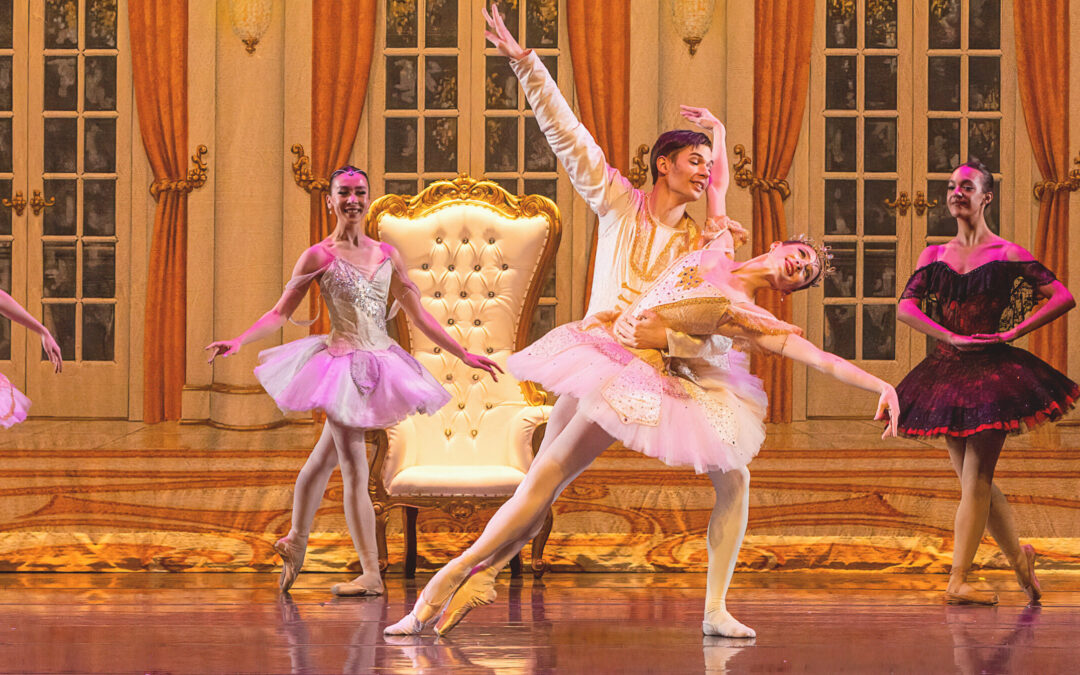 During a performance of The Nutcracker, Sage Feldges and aldrin Vendt perform together while three other female dancers watch behind them posed in b plus. Feldge, in a pink tutu with gold trim and pink tights, leans back into the crook of Vendt's left arm as he lunges to his left. Her legs are in a sus-sous position on pointe and her arms are out to the side, her right arm raised slightly higher to the left of Vendt's face. Vendt wears a white tunic and white tights and slippers.