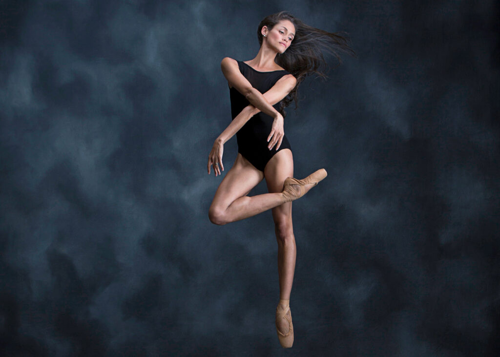 In front of a dark blue-gray marbled backdrop, a female dancer in a black leotard and nude pointe shoes jumps in a contemporary over-crossed passé position, her arms crossed at the elbows in front of each other. She looks off to the right serenely, her dark hair flying around her.