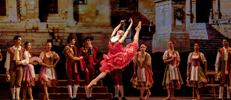 Victoria Jaiani does a large sissone with her left leg in attitude, splitting her legs and arching her upper body back with her arms in fifth position, almost kicking her head. She wears a red SPanish style dress with a knee legnth skirt and tan pointe shoes. Behind her is the set of a town square, and other dancers in various Spanish-style peasant costumes stand a watch her.