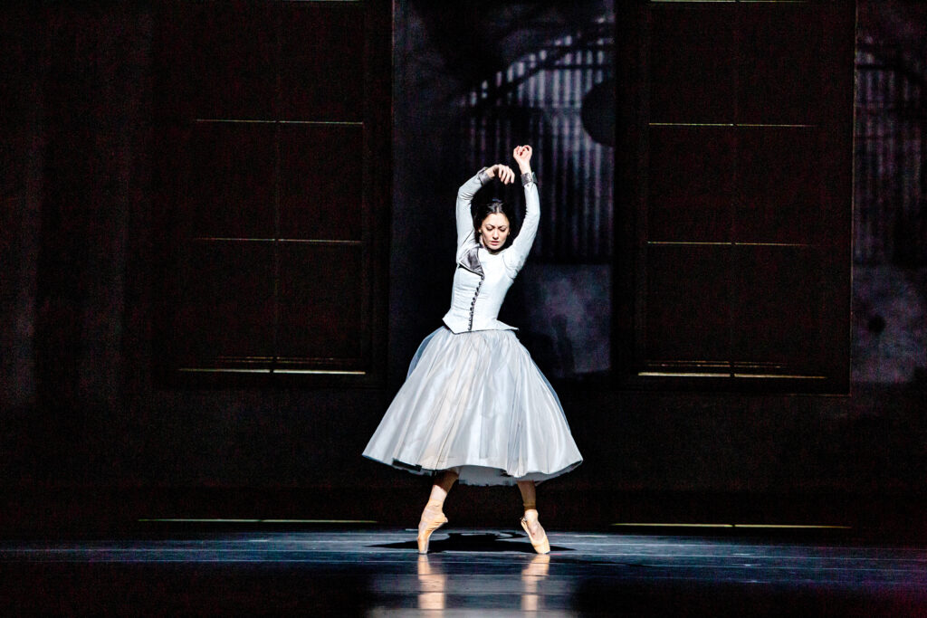 Victoria Jaiani balances on pointe in a fourth position effacé and lifts her arms up her head as she looks down to the floor. She wears a gray dress with a knee-length full tutu skirt, long sleeves and a high neck. She dances on a darkened stage.