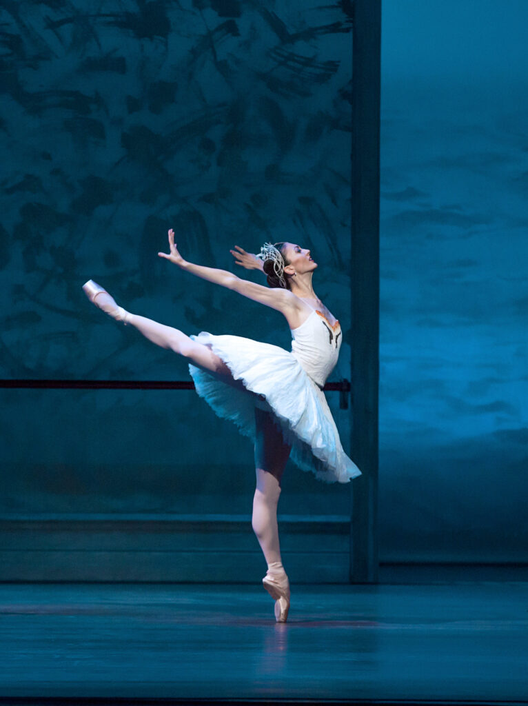 Victoria Jaiani dances onstage during a performance of Swan Lake. She does a relevé attitude derriere with her right leg lifted behind her and presses both arms back, arching her upper body. She wears pink tights, pink pointe shoes, a white tutu and a crown of feathers.