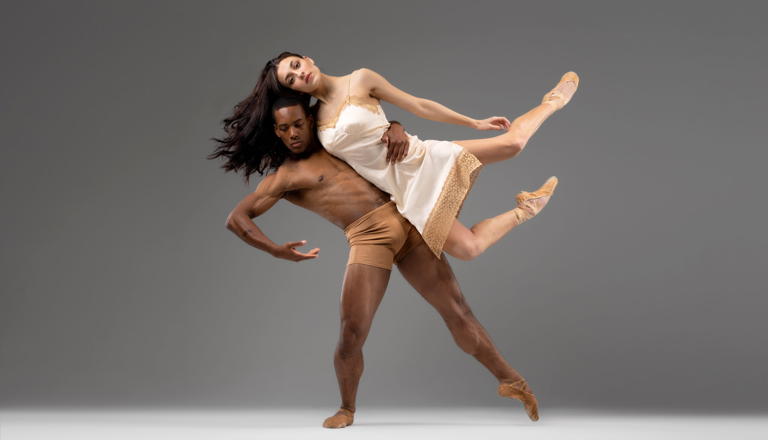 In front of a gray backdrop, a male dancer lifts a female dancer on the side of his body. She bends her legs in attitude, her hip in line with his as her legs reach on the upward diagonal. He lunges in tendu a la seconde with a stylized arm position, wrapping his left around her waist for support and mimicking the same shape with his right.