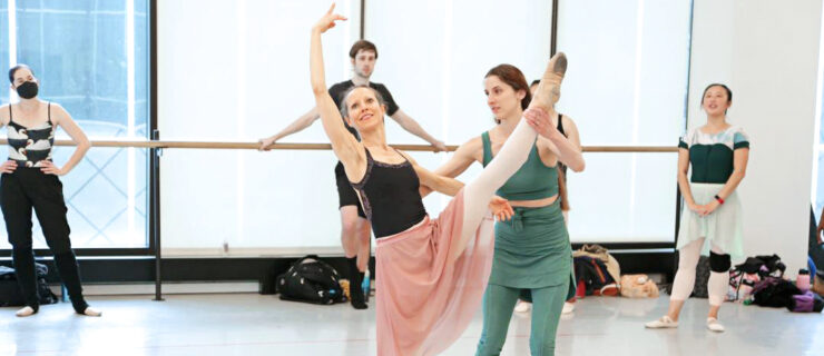 Allison DeBona gently holds the leg and lower back of an adult female ballet student as she practices a développé devant in effacé in the center of a dance studio. DeBona wears a green tank top, ballet skirt, and leggings and ballet slippers and looks toward the student, who looks up toward her lifted right arm with a smile. The student wears a black leotard, long pink skirt, footless pink tights and ballet slippers. Behind them, other dancers stand along the studio's floor-to-ceiling windows and watch.