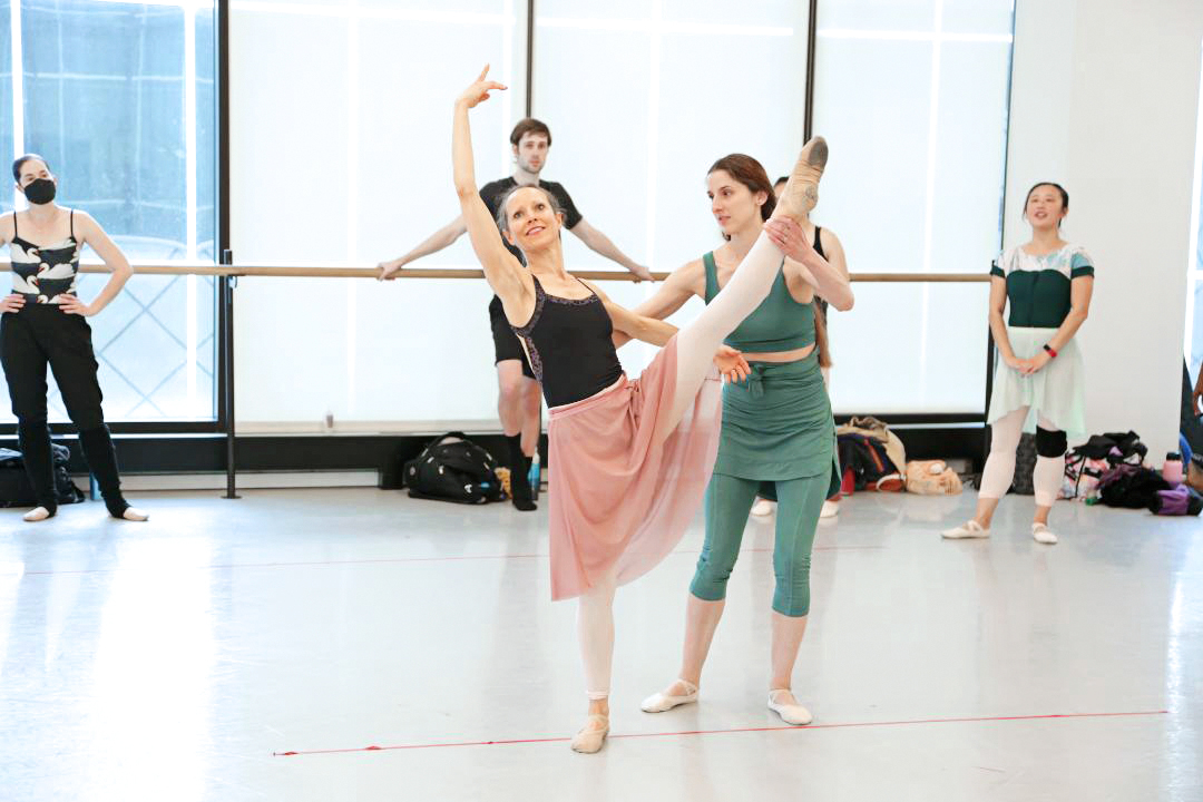 Allison DeBona gently holds the leg and lower back of an adult female ballet student as she practices a développé devant in effacé in the center of a dance studio. DeBona wears a green tank top, ballet skirt, and leggings and ballet slippers and looks toward the student, who looks up toward her lifted right arm with a smile. The student wears a black leotard, long pink skirt, footless pink tights and ballet slippers. Behind them, other dancers stand along the studio's floor-to-ceiling windows and watch.