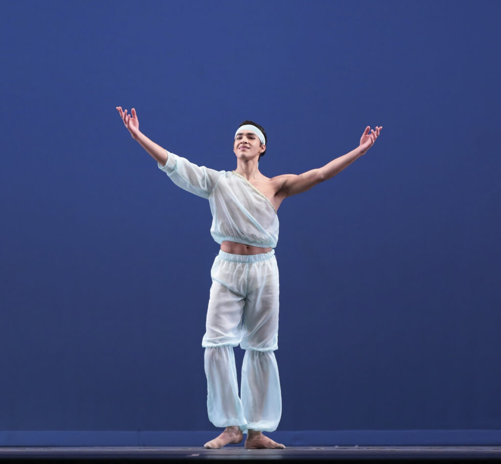 Sergio Suarez stands center stage in front of a blue backdrop and raises his arms up to take a bow. He wears filmy blue harem pants and an asymmetrical top with one elbow-length sleeve. He wears a blue matching headband around his forehead and smiles broadly.