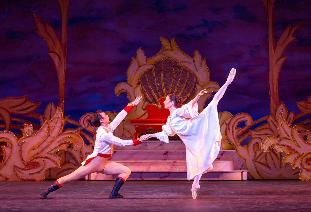 Kevin Gael Thomas and Sarah Tryon dance together during a performance of The Nutcracker. Gael Thomas, wearing a princely white jacket with red and gold trim, tan tights and black boots, lunges on his left leg in profile, facing stage left, and holds Tryon's hand with his right hand as she does a penché facing him on pointe. Tryon wears a long white nightgown with puffy sleeves. The dance in front of a small marble staircase and throne