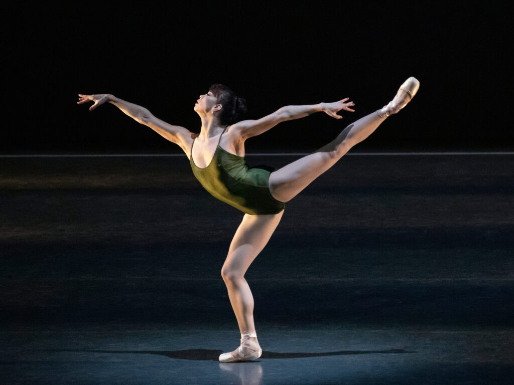 Onstage, Georgina Pazcoguin wears a deep olive-colored leotard and pointe shoes. She does a lunging arabesque on flat, her arms in a wide first arabesque position.