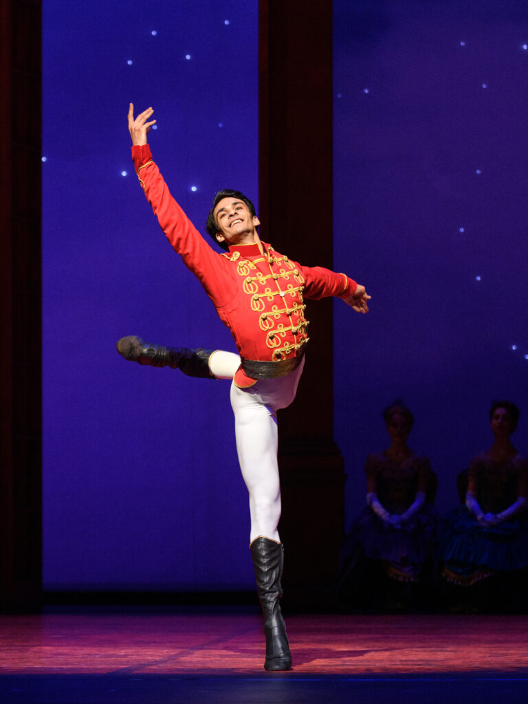 Constantine Allen, portraying a prince onstage during a performance, is shonw finishing a renversé, his left leg in attitude and his right arm lifted high and his left out to the side. He smiles triumphantly, arching his upper body slightly to the right. He wears a red jacket with gold buttons, white tights and black boots. Behind him, the set shows a starry night.