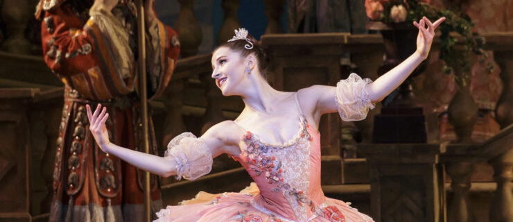Indiana Woodward performs onstage as Princess Aurora in The Sleeping Beauty. She is shown from the thigh up wearing a pink tutu with rosette embellishments on the bodice and tutu and puffy sleevelets on her upper arms. She neds her body slightly to her right and looks over her right shoulder, smiling brightly, and opens her arms generously, curving them up at the elbow.