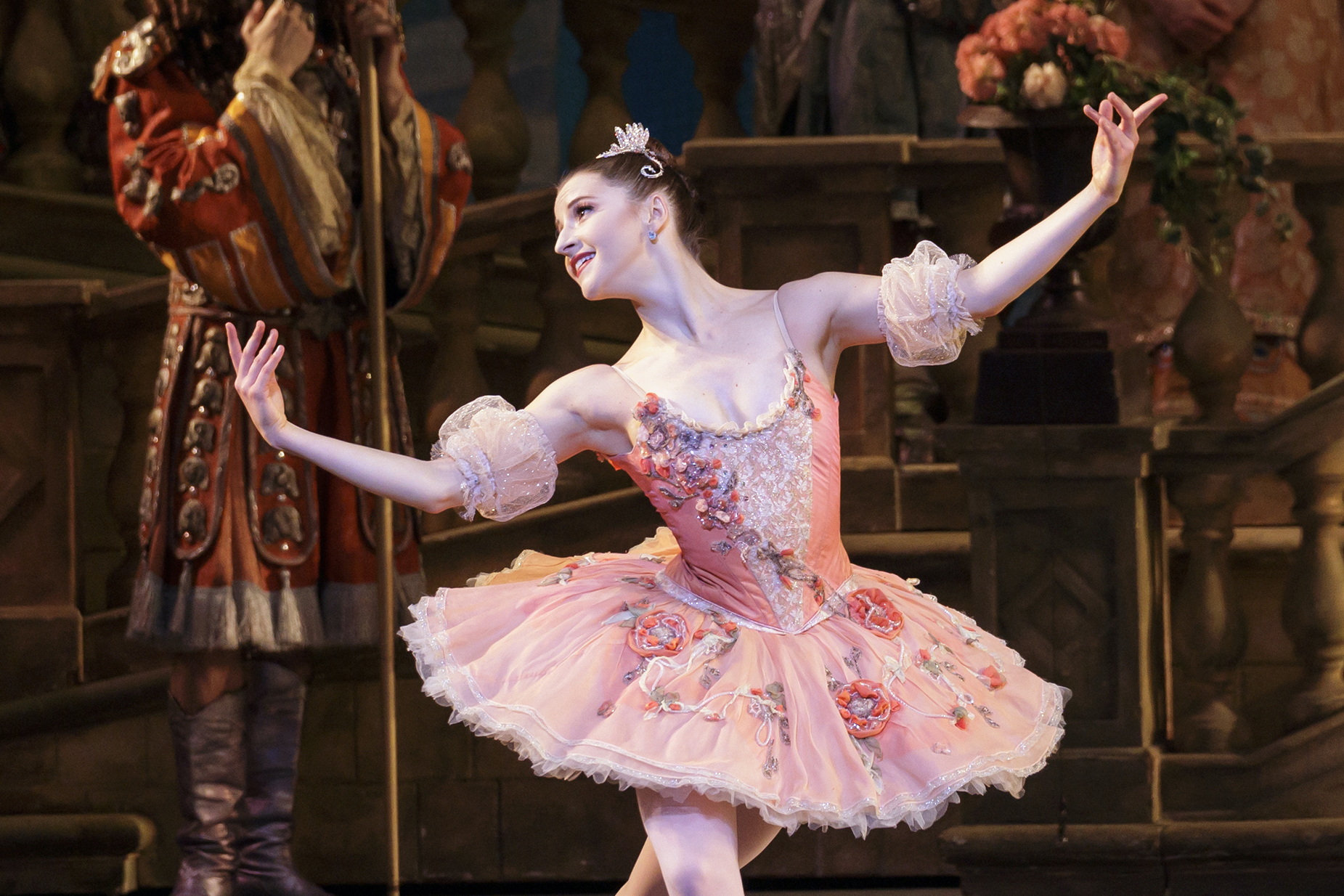 Indiana Woodward performs onstage as Princess Aurora in The Sleeping Beauty. She is shown from the thigh up wearing a pink tutu with rosette embellishments on the bodice and tutu and puffy sleevelets on her upper arms. She neds her body slightly to her right and looks over her right shoulder, smiling brightly, and opens her arms generously, curving them up at the elbow.