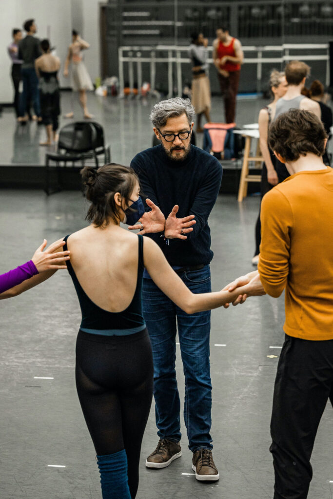 In a large dance studio, Paul Vasterling stands in front of a group of dancers and gives notes to them, placing his wrists together, palms facing out and fingers splayed. He wears a dark blue turtleneck sweater, jeans and brown sneakers with white soles. A famale dancer and male dancer face him, joining hands in a pose, and listen. The woman wears a black low-back leotard and tights, while the male dancer wears a gold long-sleeved shirt and black tights.