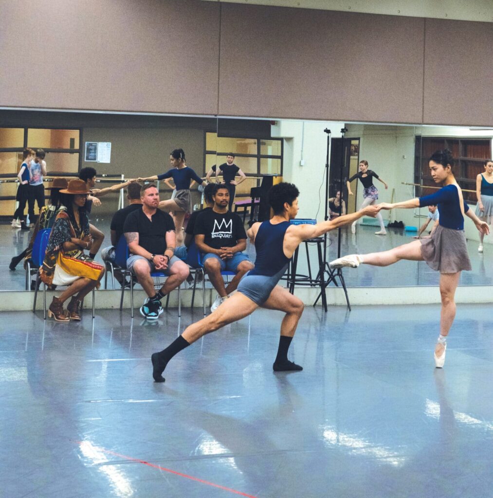 A male and a female dancer rehearse a pas de deux in a mirrored studio as three people watch at the front of the room, including Katlyn Addison (far left). The female dancer, on pointe, extends her right leg devant and holds hands with her partner, who reaches to her in a deep lunge.