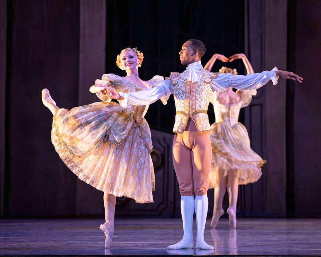 Loren Walton is supporting Rylee Ann Rogers in an attitude on pointe. They are wearing period costumes from "The Glass Slipper." Walton is in first position, with his left arm extended in second position allongé, as he looks toward his partner over his right shoulder. 