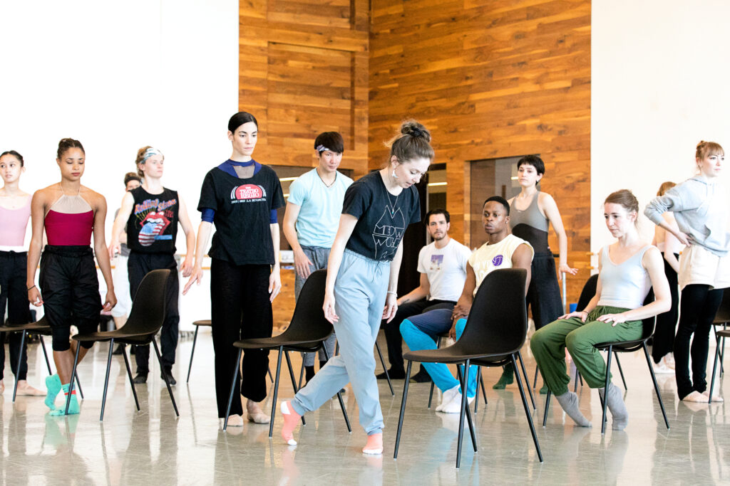 Melody Mennite, wearing a black T-shirt, gray sweatpants and pink socks, stands with her arms at her sides and her left foot in B plus and does a slight bow with her upper body. A group of dancers stand behind her, wacthing, while another group sits in rows chairs to the left side of her. They work in a huge, airy dance studio with white walls that include wood paneling in the corner.