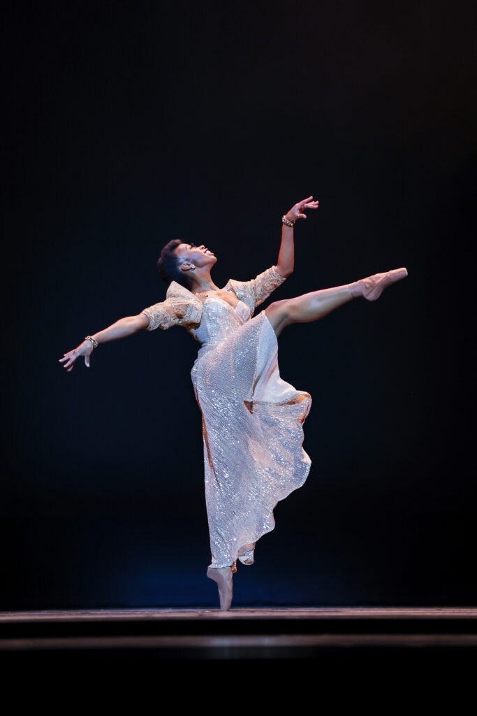 In a long, flowing white dress, Daphne Marcelle Lee does a piqué on pointe, her right leg lifted in croisé attitude devant. Her arms float at shoulder height, and she looks up at the sky.