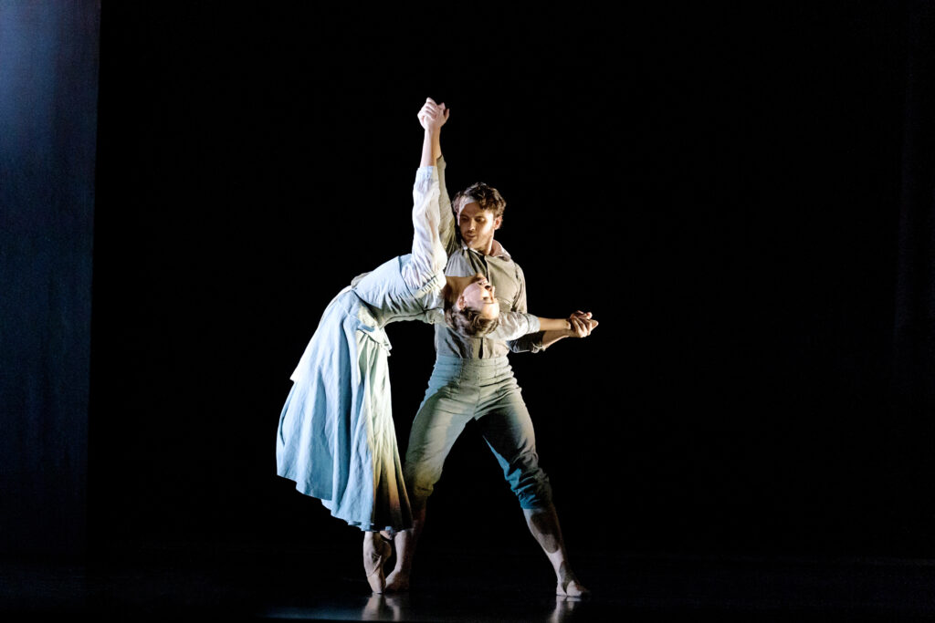 On a dark stage, a man and a woman perform an emotional pas de deux in Puritanical costumes. The woman does a sous-sus on pointe, arching backward, as the man stands behind her and holds her hands above and behind her to support her.