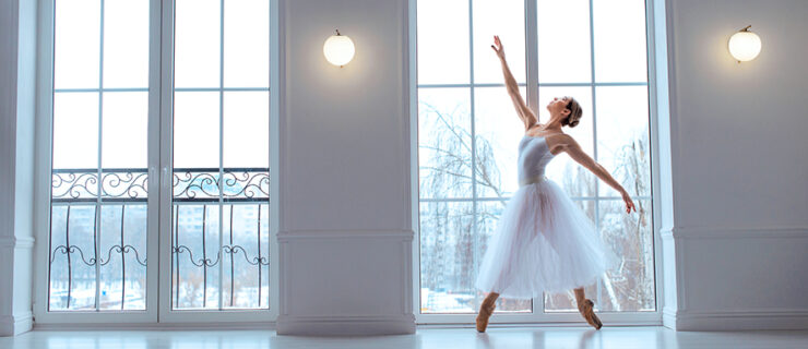 A dancer in a long white tutu stands on pointe in a wide croisé fourth position. She poses in a large, floor-to-ceiling window in a large white dance studio and reaches her right arm high. Outside the window are snow-covered trees.