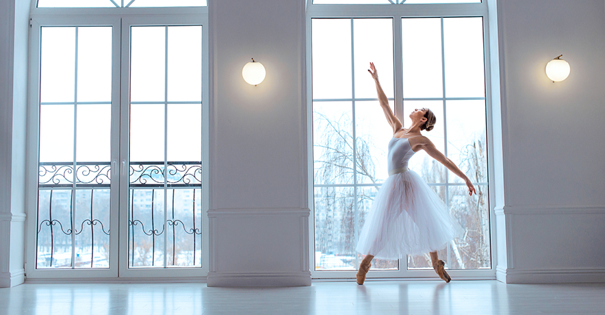 A dancer in a long white tutu stands on pointe in a wide croisé fourth position. She poses in a large, floor-to-ceiling window in a large white dance studio and reaches her right arm high. Outside the window are snow-covered trees.